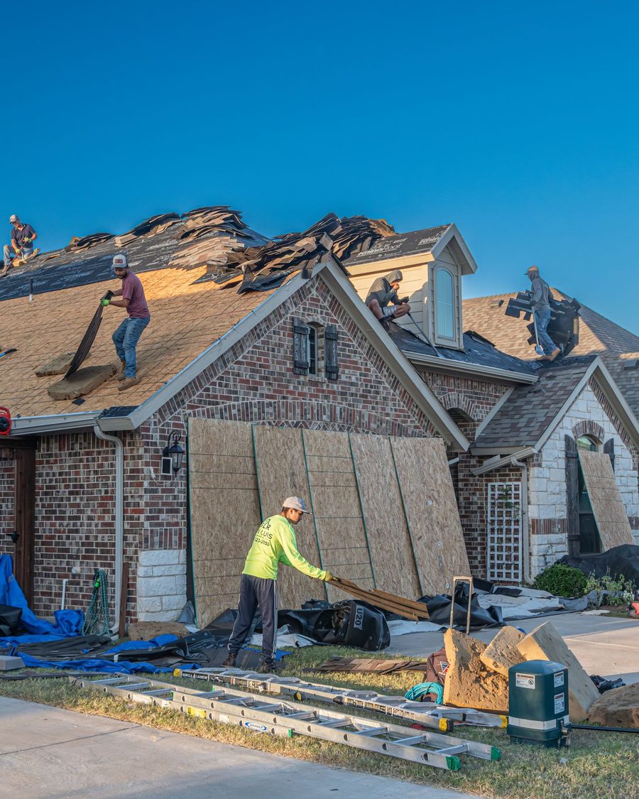 Roofers tearing off an old roof on a brick house, with a worker on the ground